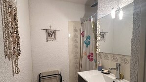 Ein Badezimmer mit weißem Waschbecken, dunklem Schrank und einem Spiegel mit Beleuchtung. Ein Blumenmuster-Duschvorhang und ein Wandregal mit einem kleinen Dekorationsobjekt sind ebenfalls vorhanden.
