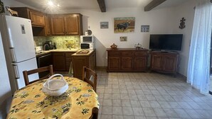 Una cuina compacta amb armaris de fusta, una taula de dinar amb mantell floral, un frigorífic, un microones i una televisió.