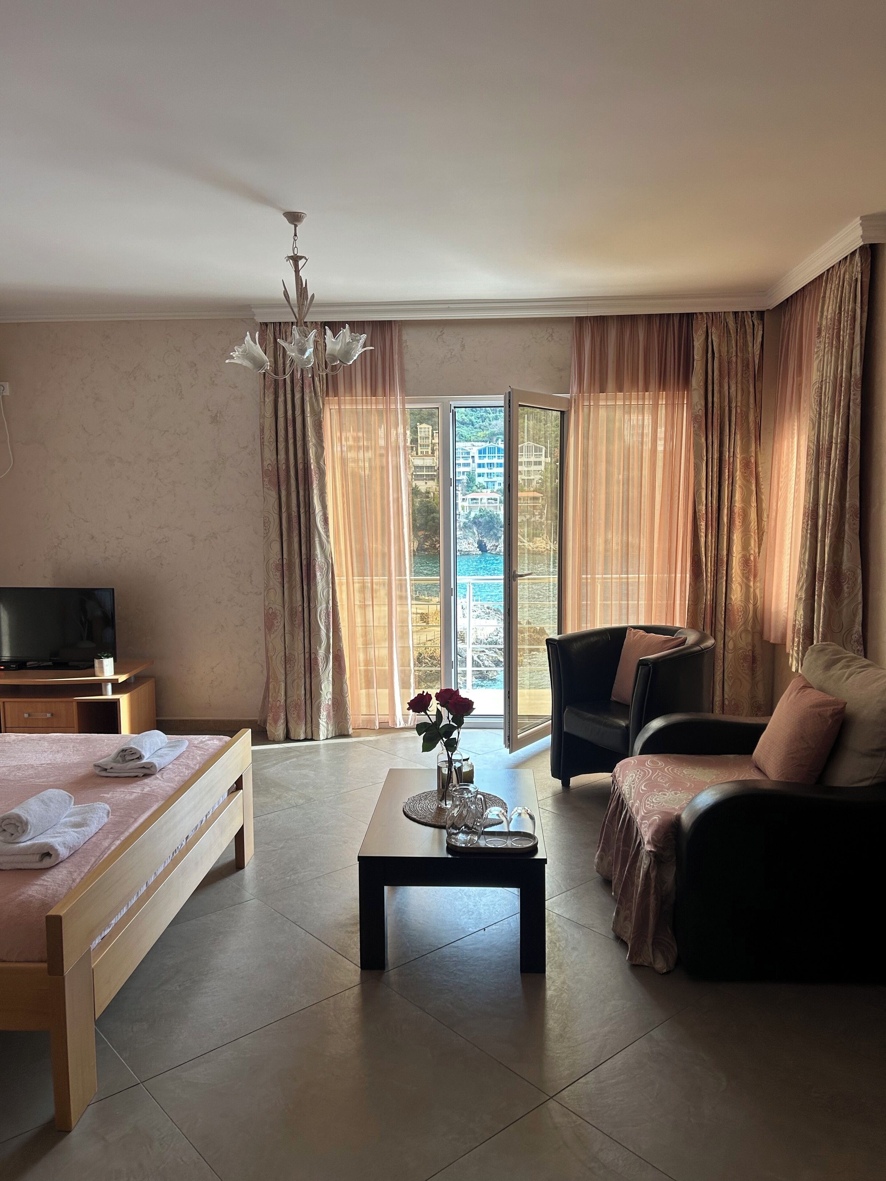 A hotel room with a bed, a sofa, a coffee table with a vase of flowers, a television, and a view of the sea through large windows.