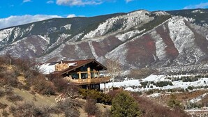 Una cabaña de madera con balcón, rodeada de montañas nevadas y un cielo despejado.
