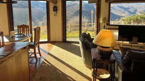 Ein großzügiger Wohnraum mit einem großen Fenster, das einen Blick auf die Berge bietet, einem Holzesstisch und einer gemütlichen Sitzecke mit einer Couch und Sesseln.