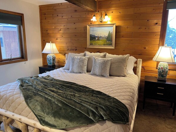 A bedroom with a wooden wall, a bed with a green blanket, two bedside tables with lamps, and a painting above the bed.