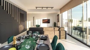 Een moderne eetruimte met een lange tafel gedekt voor vier personen, een bank op de achtergrond en een groot raam met uitzicht op de omgeving.