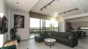 Een moderne woonkamer met een hoekbank, een ronde salontafel en een groot raam met uitzicht op de omgeving.