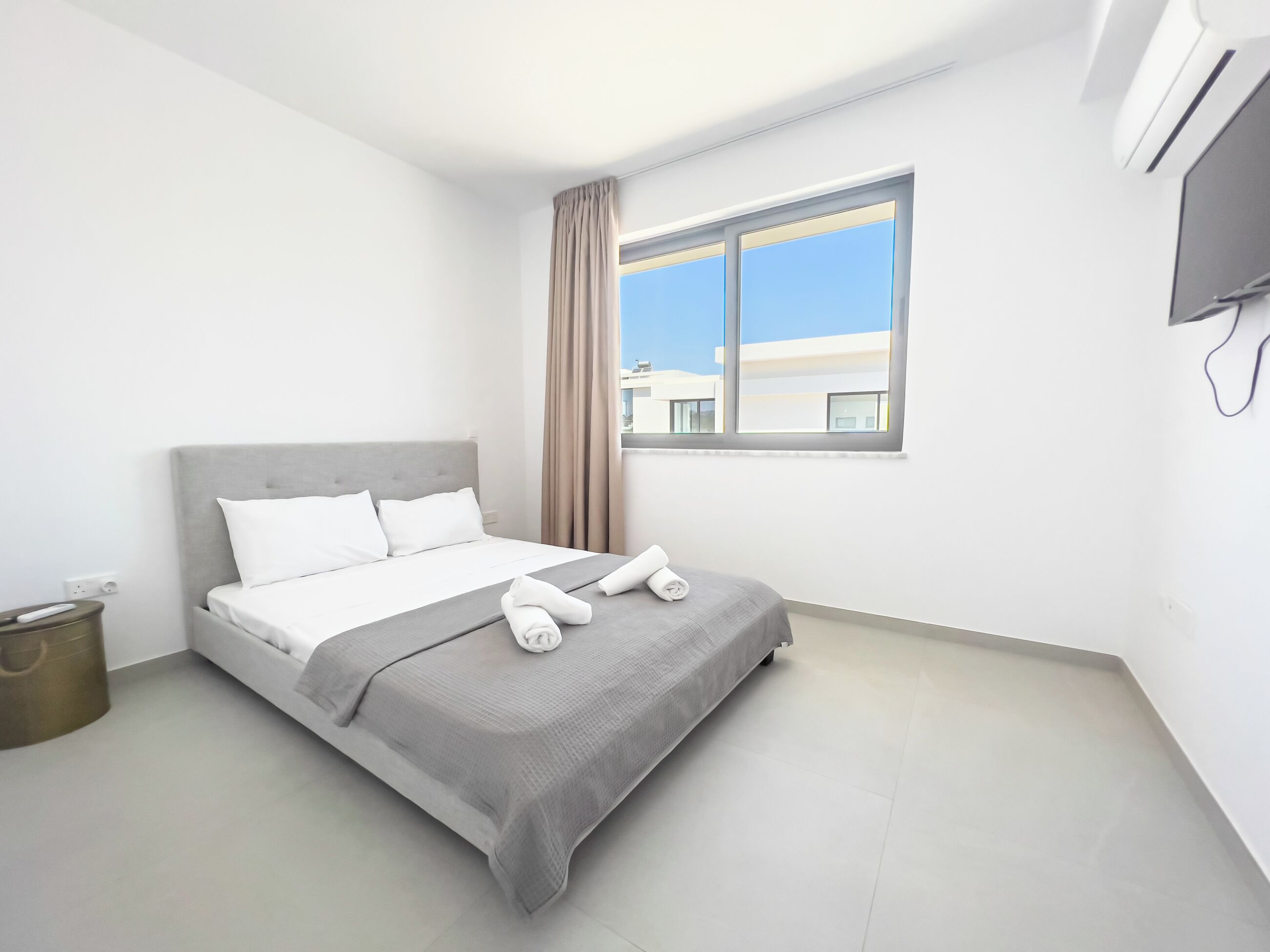 Une chambre d’hôtel moderne avec un grand lit, une fenêtre avec des rideaux, un téléviseur à écran plat fixé au mur et une petite poubelle.