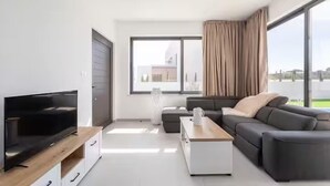Een moderne woonkamer met een flatscreen televisie, een bank, een salontafel en een groot raam met gordijnen.