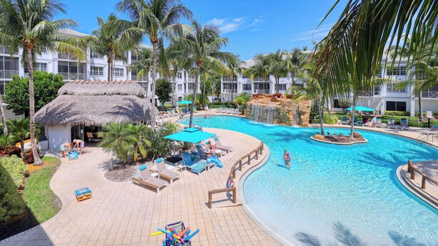 A resort pool area with lounge chairs, palm trees, and a thatched-roof hut.