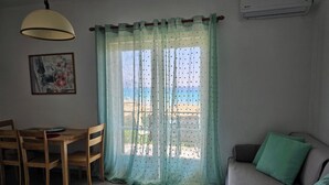 Un salon comprenant une table à manger en bois, des chaises, un canapé avec des coussins verts, une lampe suspendue au plafond et une fenêtre ornée de rideaux légers.
