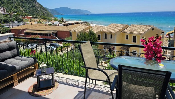 Un balcon aménagé avec un canapé, des chaises et une table, offrant une vue sur une ville côtière, avec ses bâtiments, et une vue dégagée sur la mer.