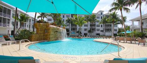 A pool area with lounge chairs, a turquoise umbrella, and a waterfall feature.