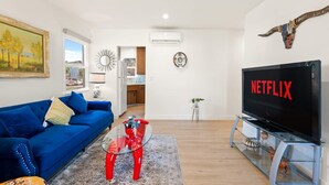 Uma sala de estar com um sofá azul, uma mesa de centro de vidro, uma TV exibindo 'NETFLIX' e uma cozinha ao fundo.