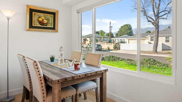 Uma sala de jantar com mesa de madeira, cadeiras acolchoadas, uma obra de arte emoldurada e vista para uma rua residencial através de grandes janelas.