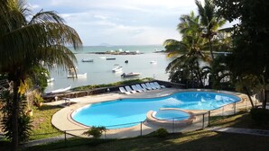Piscina con sillones de descanso, rodeada de palmeras y vista a barcos en el agua.