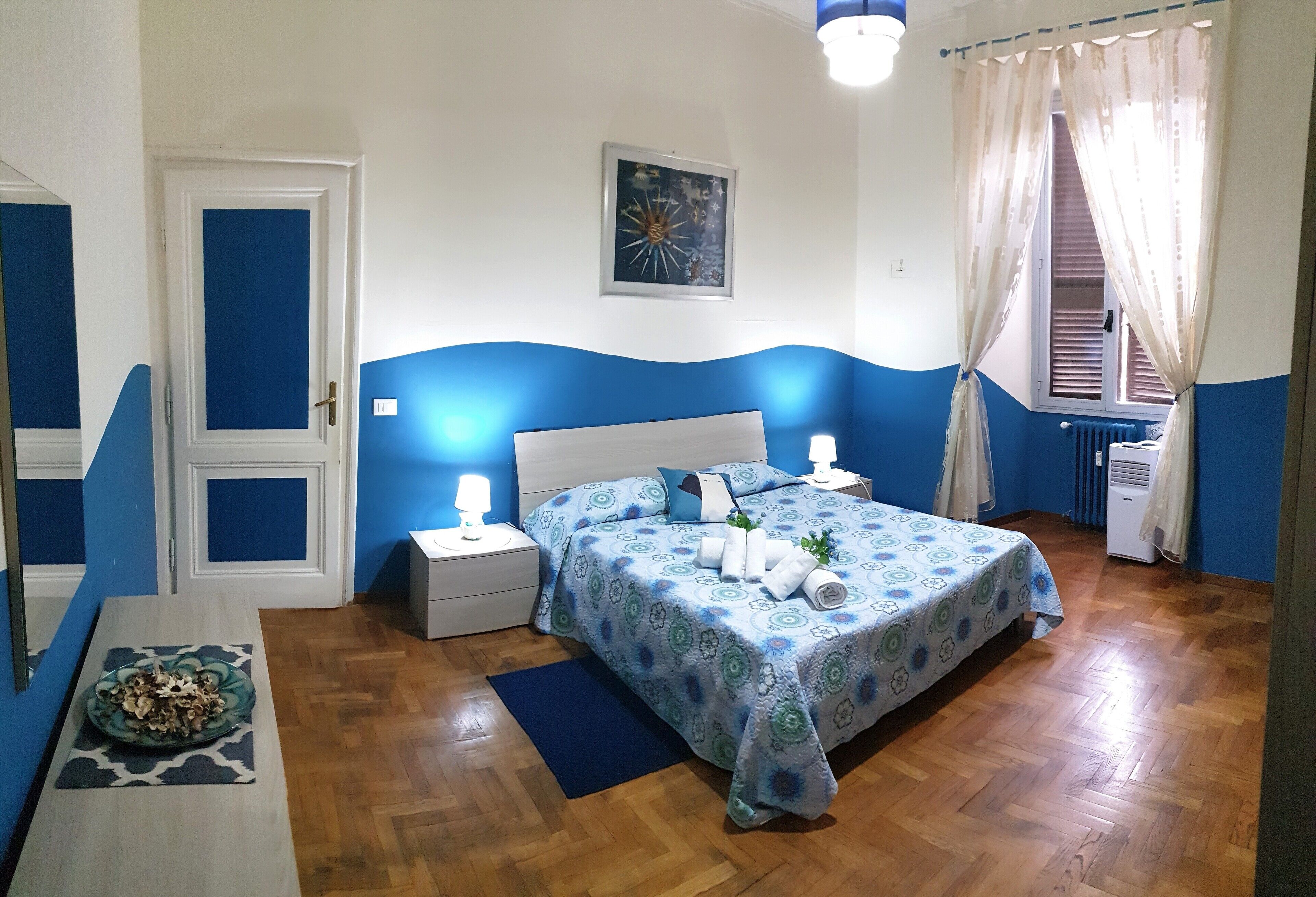A bedroom with a bed, nightstands, a mirror, a radiator, and a window with curtains.
