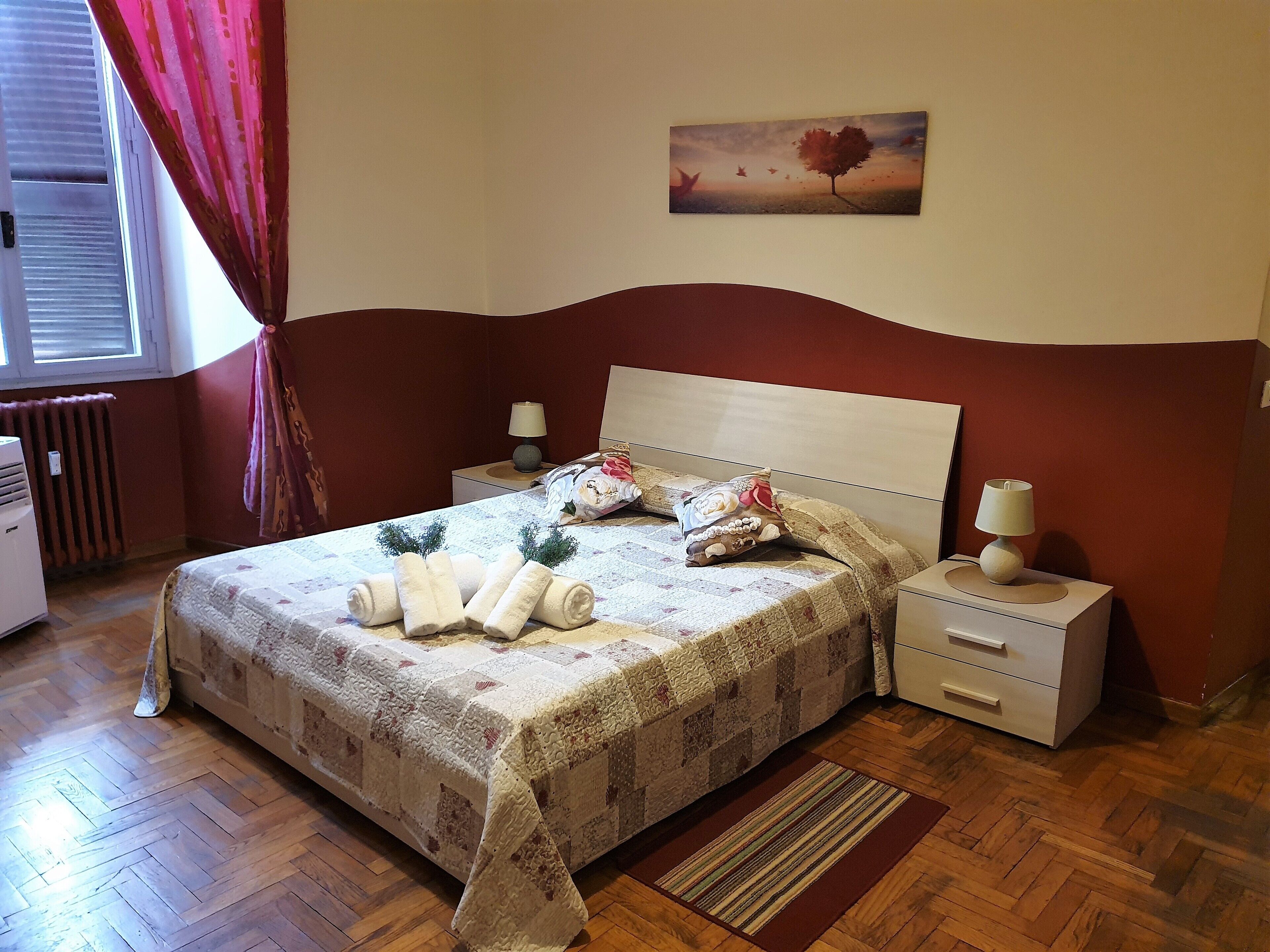 A neatly made bed with rolled towels, a bedside lamp, and a floral pillow. There is a painting on the wall above the bed and a radiator in the corner.