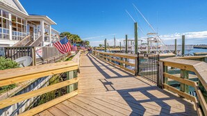 A wooden boardwalk with American flags, leading to a marina with boats and a building with a sign that reads "Open Daily at Noon." - Starfish Retreat 3/2 By Beach Star (Murrells Inlet)