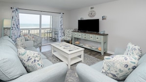 A living room with a sofa set, a coffee table, a TV, and a view of the ocean through large windows.