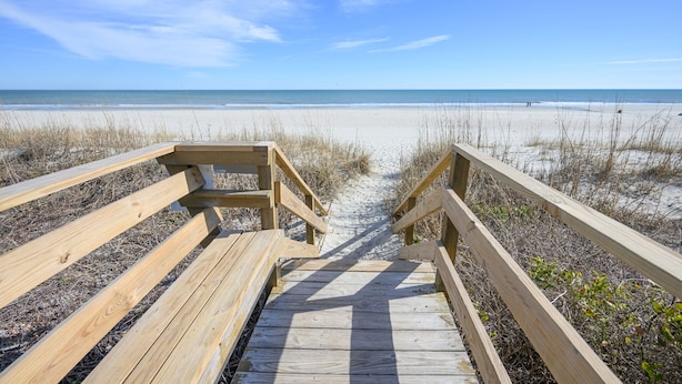 A wooden boardwalk leading to a beach with sand and sparse vegetation.