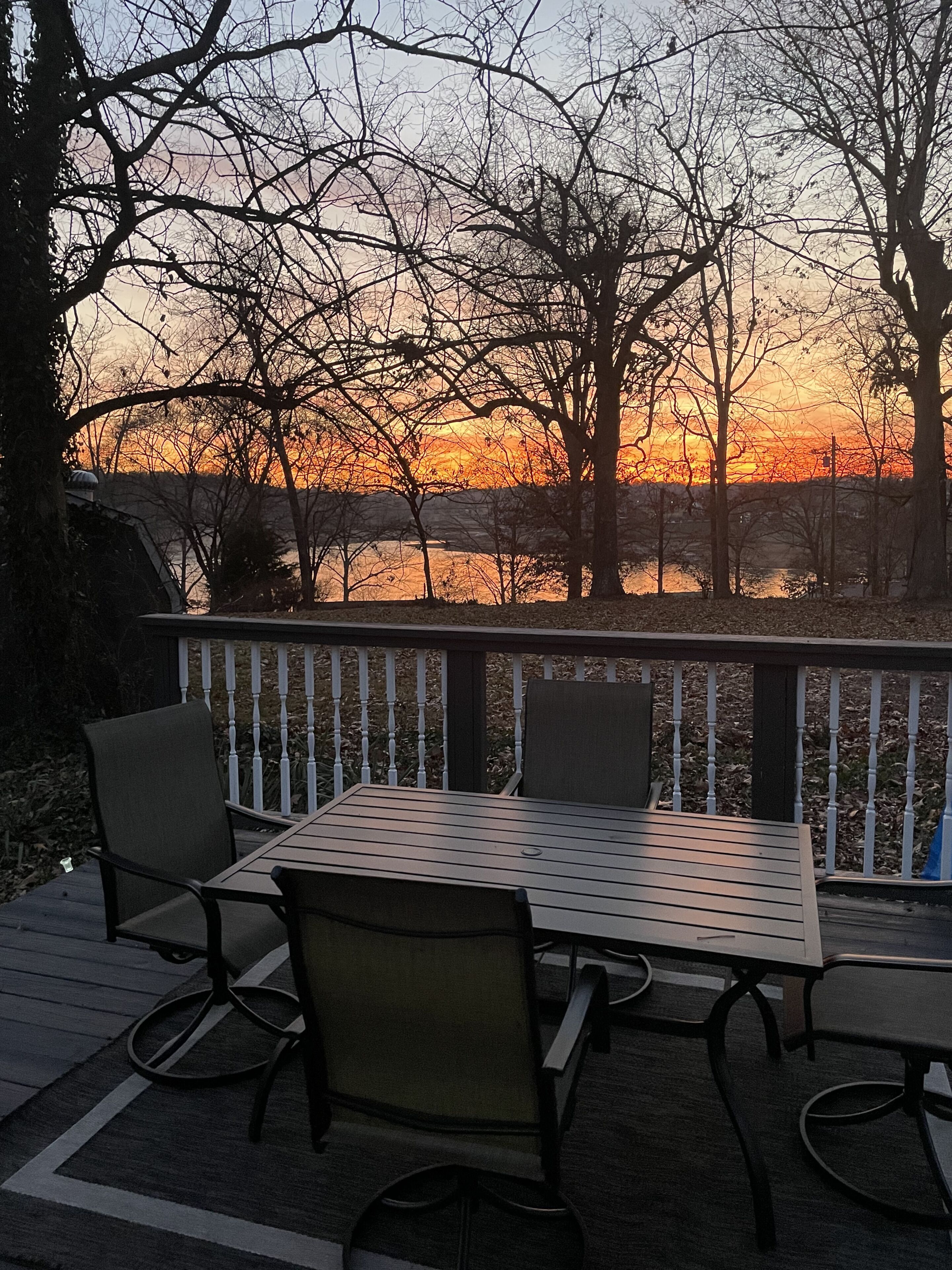 Eine Terrasse mit Tisch und Stühlen, mit Blick auf ein Gewässer und Bäume bei Sonnenuntergang.