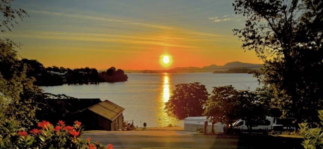 Auringonlasku vesistön yllä, kuvaan kuuluu pieni rakennus ja ajoneuvo etualalla.