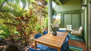 A covered outdoor dining area with a wooden table and blue chairs, surrounded by lush tropical plants and a wicker sofa. - Puamana at Princeville Island Style Townhome, Beautifully Updated, sleeps 8 (Princeville)
