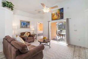 A living room with a brown sofa, a ceiling fan, a painting, and a dining area with a table and chairs.