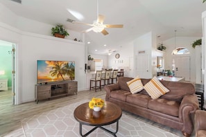 A living room with a brown leather sofa, a round coffee table with a vase of sunflowers, a ceiling fan, and a large flat-screen TV.