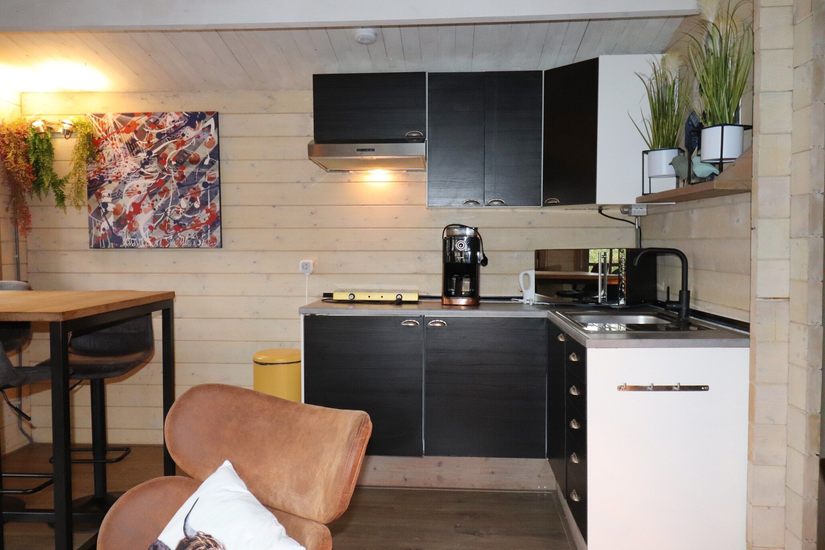 A compact kitchen with a coffee maker, a sink, and cabinets. There is a dining table and a chair in the adjacent area.