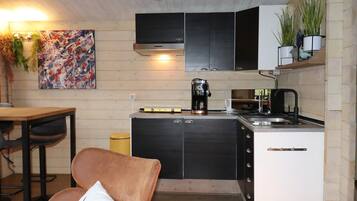 A compact kitchen with a coffee maker, a sink, and cabinets. There is a dining table and a chair in the adjacent area.