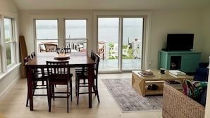 A spacious living area with a dining table, chairs, a sofa, and a television. There are skylights and large windows providing views of the outdoors. - Charming Waterfront Island Bungalow with DOCK in Narragansett! (Narragansett)