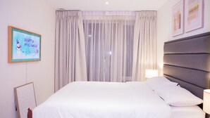Une chambre d’hôtel avec un grand lit, une tête de lit, une table de chevet avec une lampe et des rideaux légers.