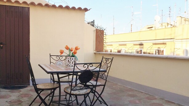 A patio with a table and chairs, a vase with flowers, and a view of buildings in the background. - Casa Tua sul tetto di Roma (Rome)