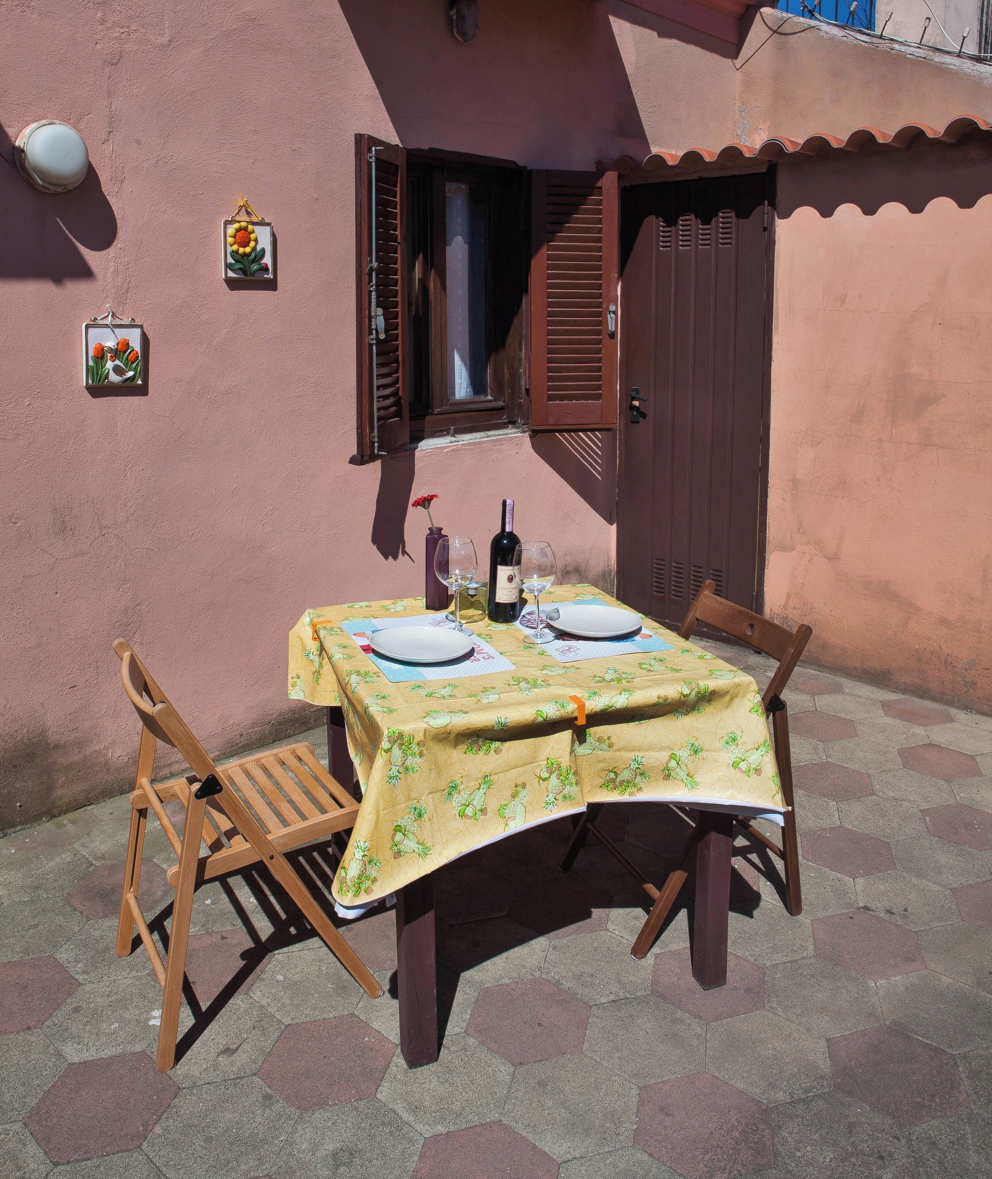 An outdoor table set with plates, wine, and glasses, a chair, and a bottle of wine on a tablecloth with a floral pattern.