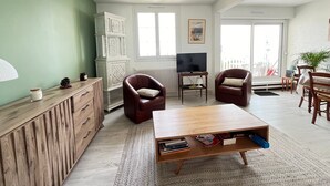 Ein geräumiges Wohnzimmer mit einem Holztisch, zwei Ledersesseln, einem Fernseher auf einem Schrank und einem großen Fenster mit Blick ins Freie.