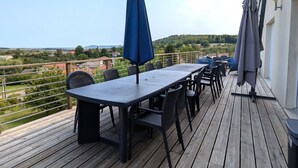 A wooden deck with a table and chairs, a blue umbrella, and a view of a green landscape.