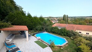 A swimming pool with a tiled deck, a covered seating area with chairs, and a view of a rural landscape.