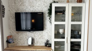 A modern living room with a flat-screen TV mounted on the wall, a wooden console table with a mirror and decorative items, a glass cabinet with dishes and glasses, and a yellow ottoman. - Renovated T2 with WiFi and parking 200m from the Spa (La Roche-Posay)