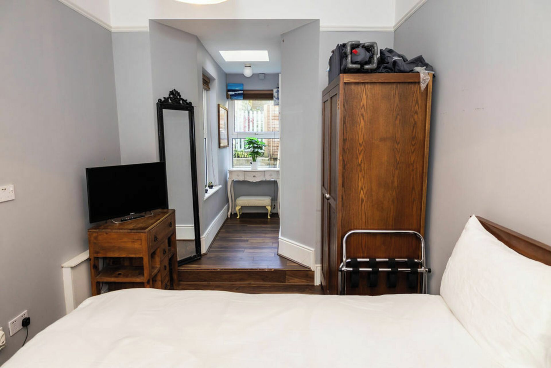 A bedroom with a bed, a wooden wardrobe, a television on a wooden dresser, and a hallway leading to a bathroom.