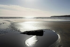 A beach with a large rock in the water, waves, and a distant cliff.
