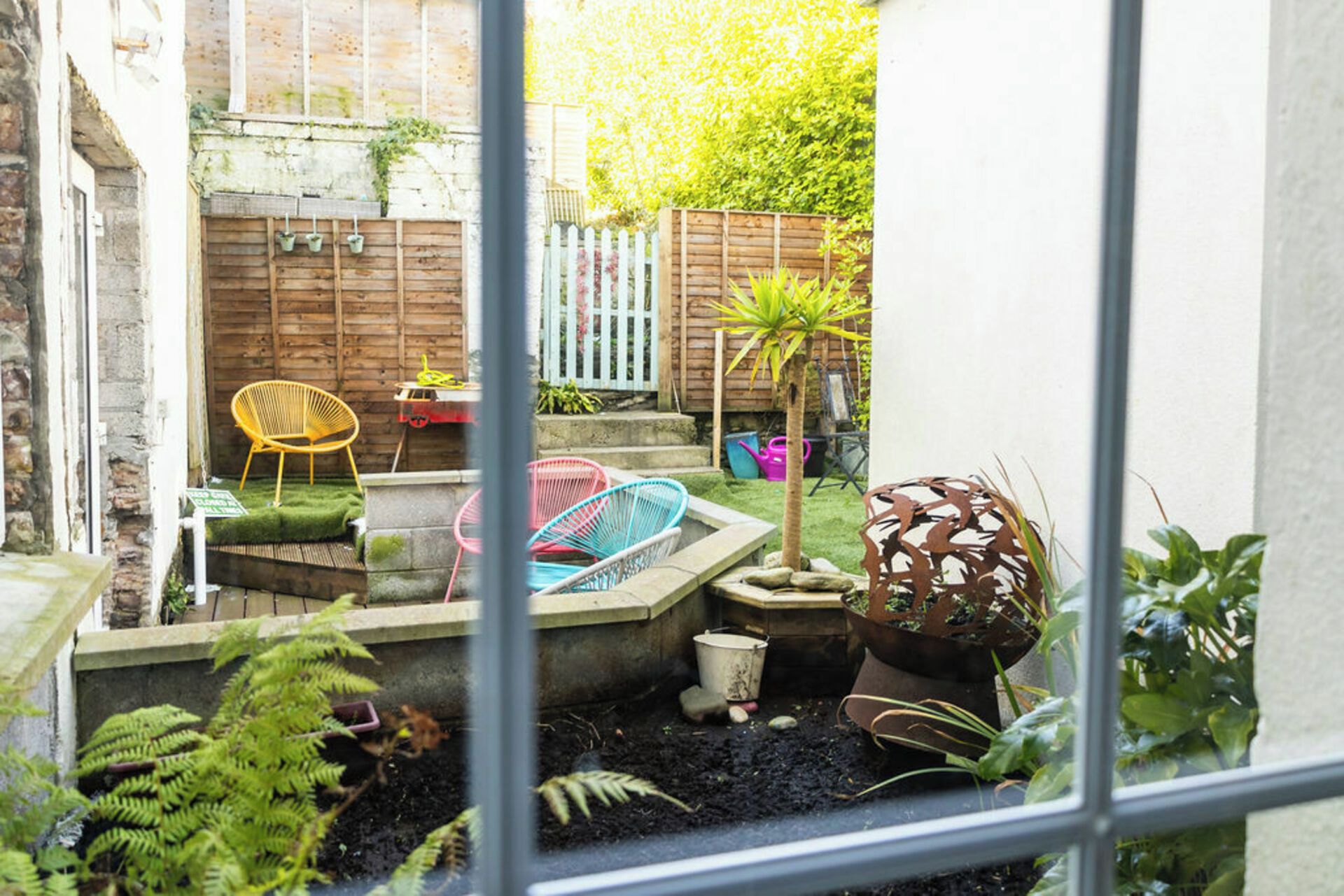 A small garden with a wooden fence, a yellow chair, a blue chair, and various potted plants.