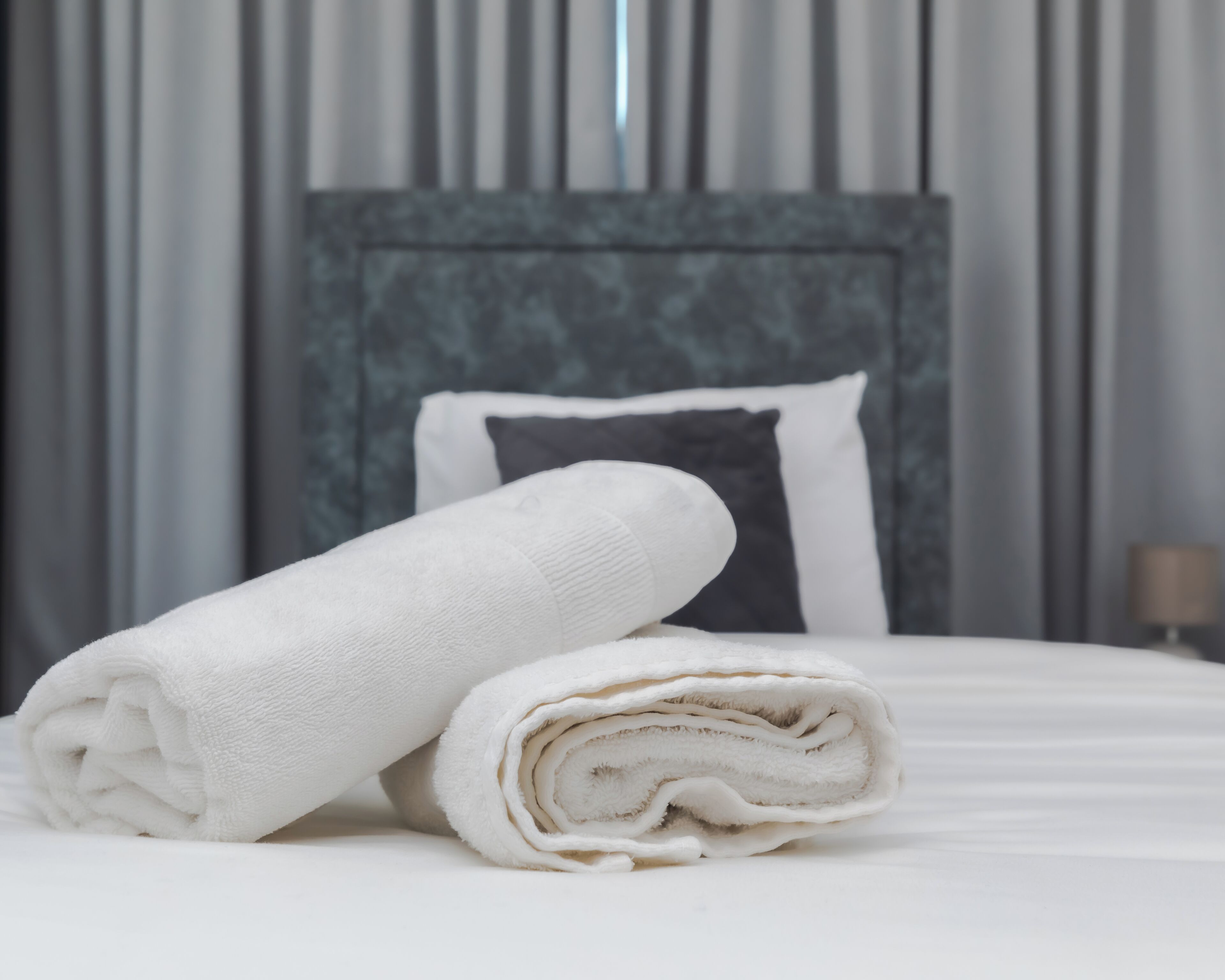 A neatly made bed with a rolled white towel and a pillow, set against a modern headboard and curtains.