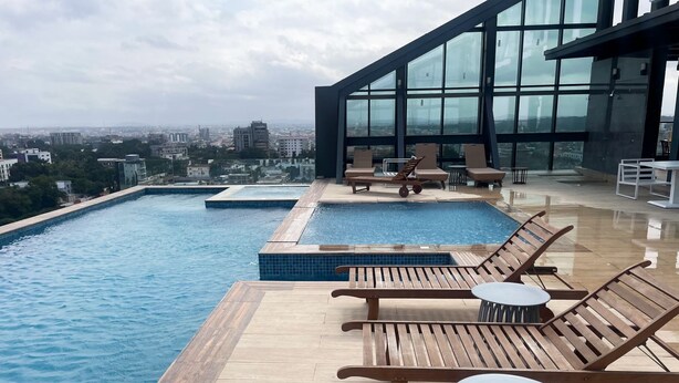 A rooftop infinity pool with wooden loungers and tables, surrounded by glass walls offering a city view. - Kass Towers (Accra)