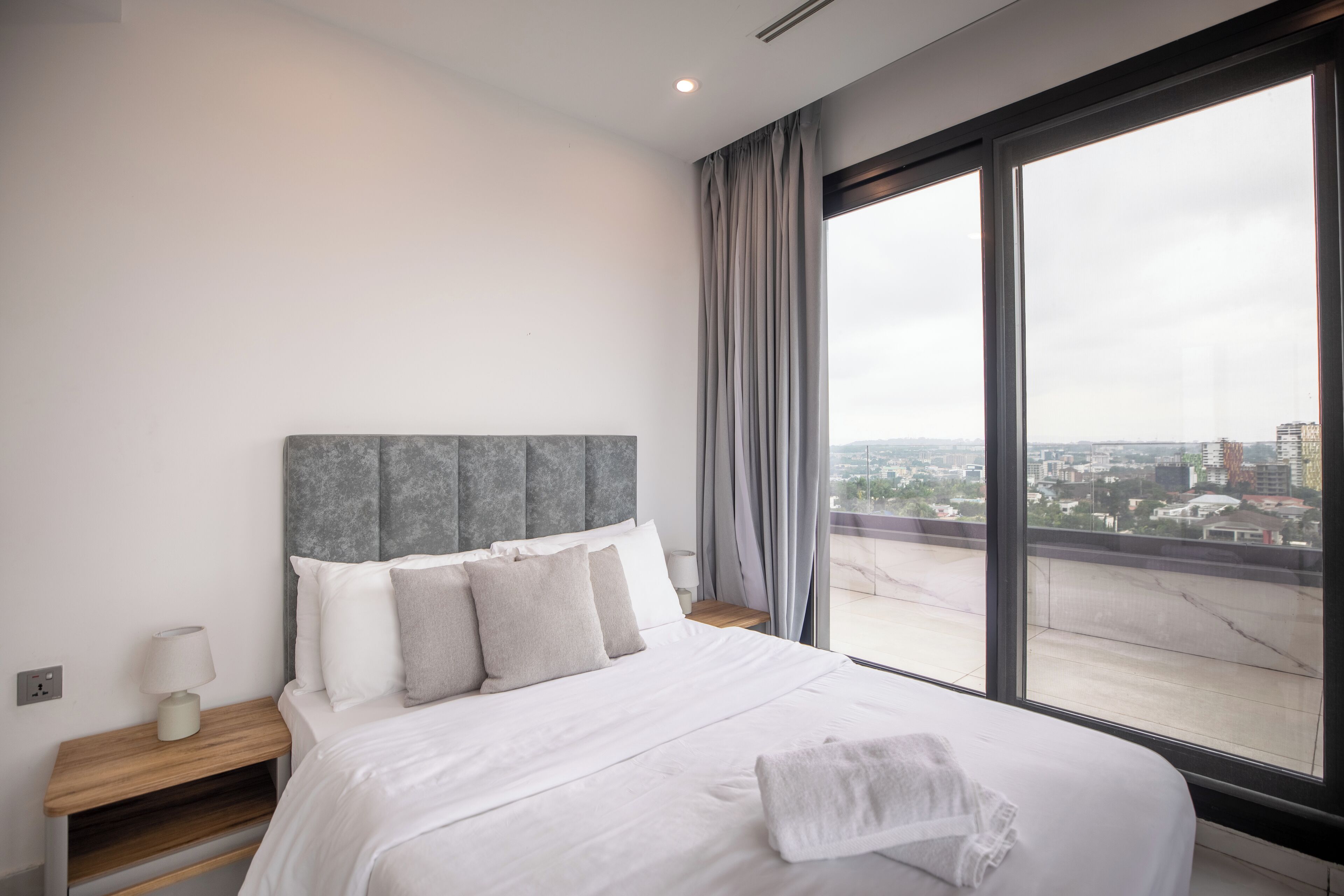 Ein modernes Hotelzimmer mit einem großen Bett, einem Holfnachtkästchen, einem Nachttisch mit Lampe und Blick auf die Stadt durch große Fenster.