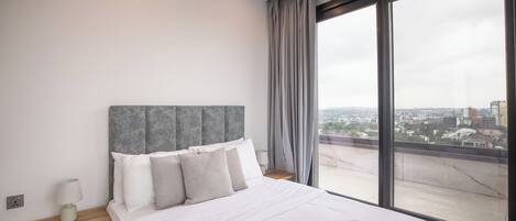 A modern hotel room with a large bed, a wooden nightstand, a bedside table with a lamp, and a view of the city through large windows.