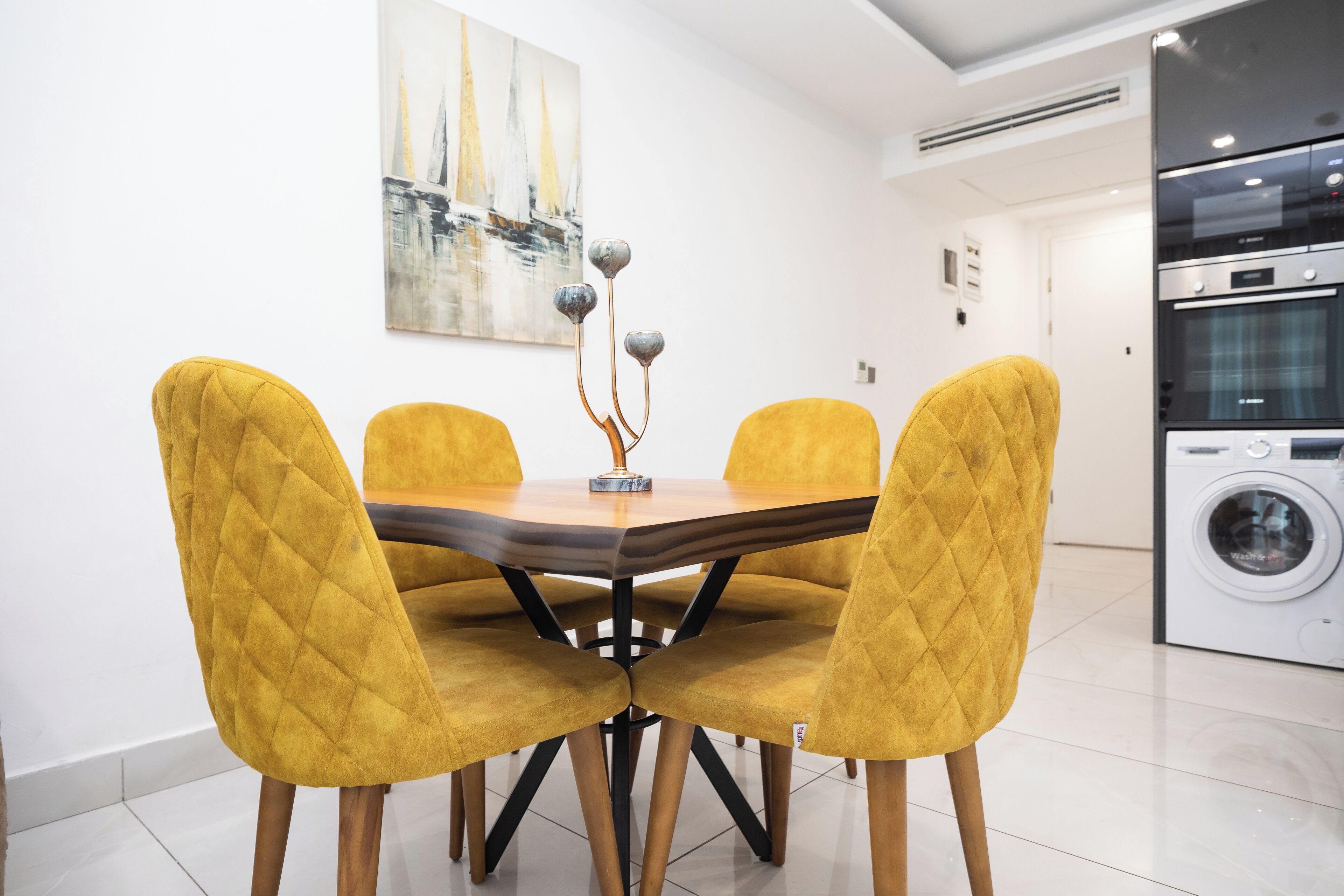 Un'area pranzo con un tavolo rotondo e quattro sedie gialle, un quadro al muro e una lavatrice sullo sfondo.