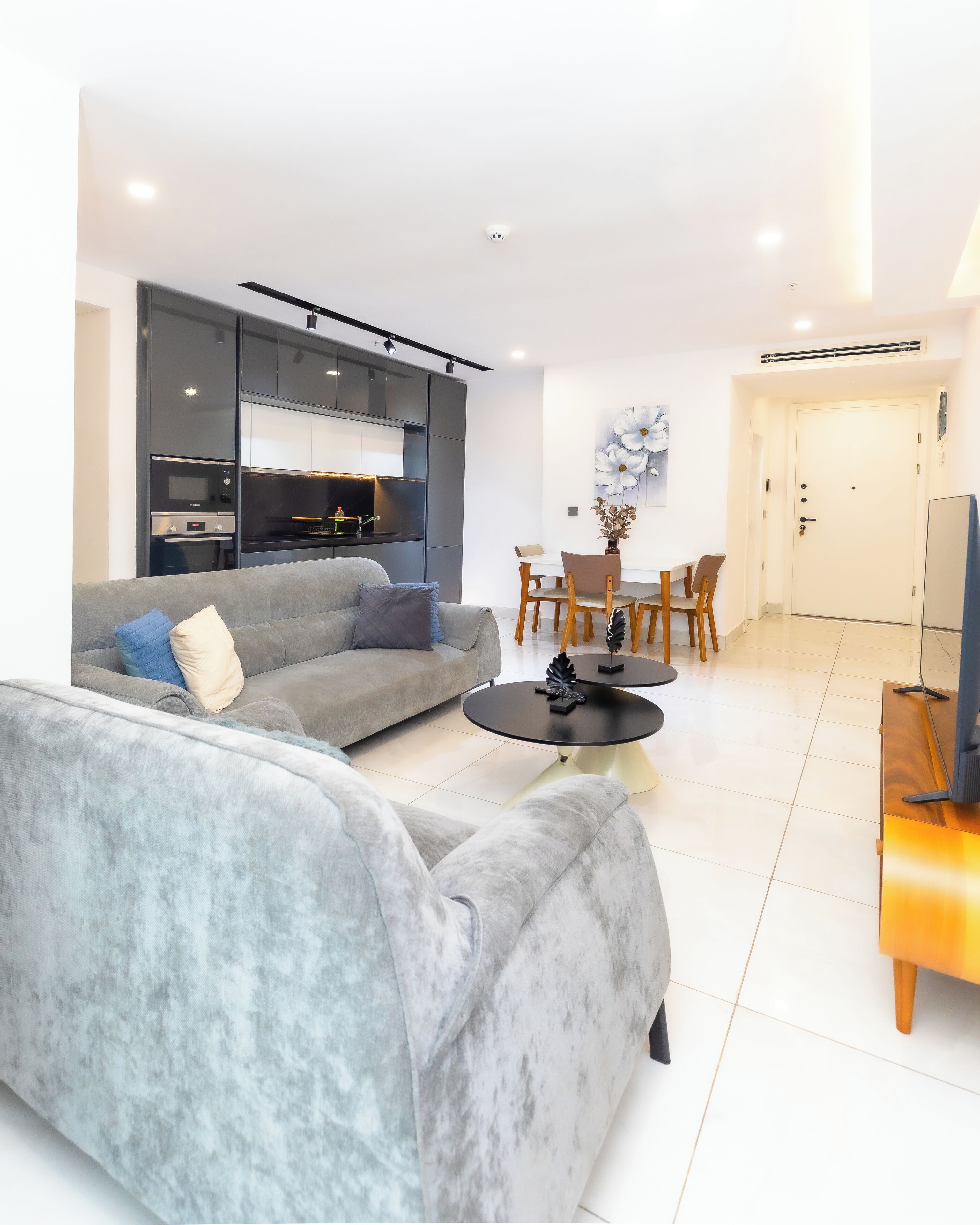 Una sala de estar moderna con un sofá gris, una zona de comedor con sillas de madera, una cocina con electrodomésticos integrados y una puerta blanca.