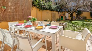 Patio con tavolo apparecchiato per mangiare, circondato da sedie bianche, con un giardino e un piccolo edificio sullo sfondo.