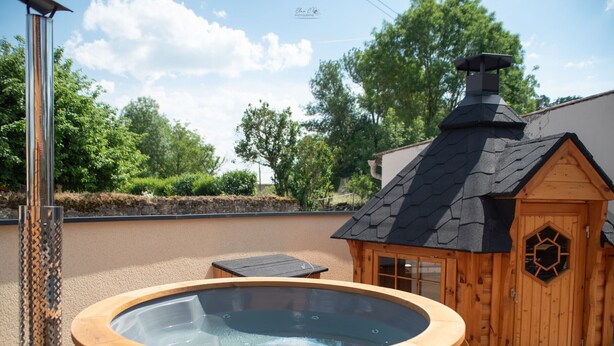 Un bain à remous en bois doté d’une fenêtre hexagonale, d’une cheminée et d’une petite structure en bois avec un toit en pente.