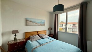 Une chambre à coucher comprenant un lit, une table de chevet, une lampe, un tableau et une fenêtre donnant sur des immeubles.
