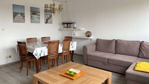 A dining area with a table set for six, surrounded by wicker chairs, a wooden coffee table with a plate of food, and a sofa in the living area. - 3-room apartment with balcony and air conditioning in Balaruc-les-Bains (Balaruc-les-Bains)